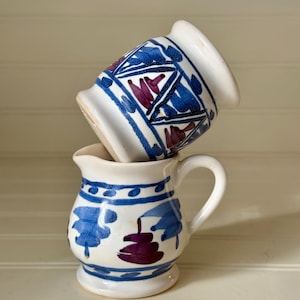 May include: Two small, white ceramic pitchers with blue and purple floral designs. One pitcher is upright, while the other is tilted on top. The pitchers have a handle and a rounded base. The design includes geometric shapes and leaf-like patterns.