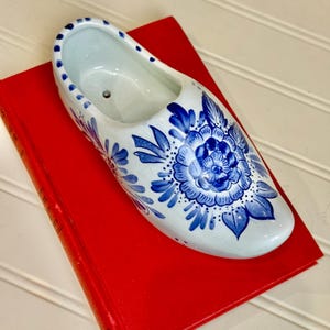May include: A white ceramic Dutch clog planter with blue floral designs. The planter is resting on a red book. The clog features a detailed blue flower and leaf pattern, typical of Delftware.