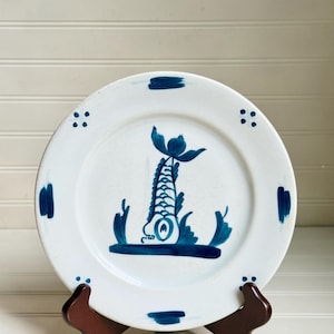 May include: White ceramic plate with a hand-painted blue fish design. The plate has a simple, elegant style with a fish in the center and blue accents around the rim. The plate is displayed on a dark brown wooden stand.