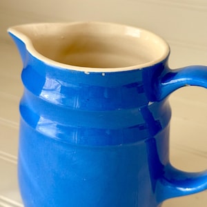 May include: A blue ceramic pitcher with a handle and a spout. The pitcher has a glossy finish and a cream-colored interior. The pitcher is a classic design, suitable for serving beverages.