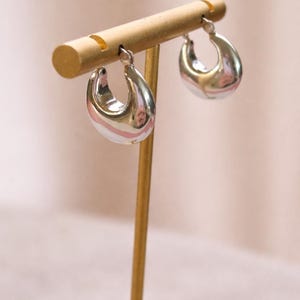 May include: A pair of silver-colored, teardrop-shaped hoop earrings displayed on a gold-colored earring stand. The earrings have a smooth, reflective surface and a rounded bottom. The stand has a cylindrical top and a thin, vertical post.