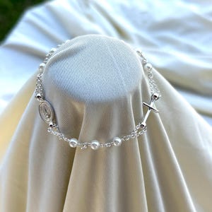 May include: A silver bracelet with white pearl beads and a silver cross charm. The bracelet also features a silver pendant with a religious image.