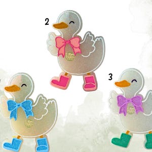 May include: Three glittery duck-shaped ornaments with bows and boots. Each duck has a different colored bow and boots: blue, pink, and purple. The ducks have orange beaks and the text "5 Star Freshies" on the bows.