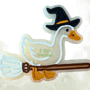 May include: A white duck with an orange beak and feet, wearing a black witch hat with an orange band, flying on a brown broomstick with a light blue brush. The text 5 Star Freshies is printed in light green on the body of the white duck.