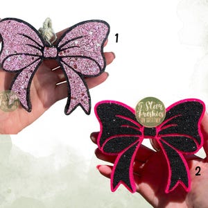 May include: Two bow-shaped air fresheners. The first is pink with silver glitter and a black outline, featuring a hanging string. The second is black with silver glitter and a bright pink outline. Both have a circular logo reading 5 Star Freshies.