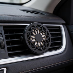 May include: A circular black car air freshener vent clip featuring a geometric floral cutout pattern with a solid center and a light-colored circular insert visible behind the black lattice frame, attached to a horizontal vehicle air vent.