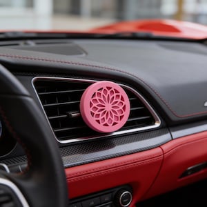 Puede incluir: Un ambientador de coche rosa y circular con un diseño floral está sujeto a una rejilla de ventilación del coche. El interior del coche presenta asientos de cuero rojo y un salpicadero negro con costuras rojas.