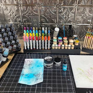 May include: A workspace with various art supplies, including a set of colorful markers, a set of ink pads, a set of paint pens, and a set of paint bottles. There is also a piece of paper with blue and white paint on it.