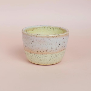 May include: A small, speckled ceramic bowl with a rounded shape. The bowl features a color gradient, transitioning from a pale yellow base to a light pink band, and a speckled white rim. The interior is a soft yellow.