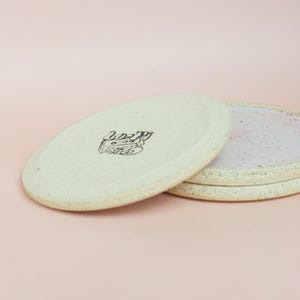 May include: A stack of three speckled ceramic plates. The top plate is off-white with a black graphic and a slightly raised edge. The plates have a minimalist design and are set against a pink background.