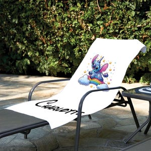 May include: White beach towel with a blue cartoon character riding a rainbow. The character has a unicorn horn and is surrounded by stars. The towel has the name "Charlotte" written on it.