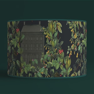 Peut inclure: Abat-jour cylindrique avec un fond sombre et un motif botanique. Le design comprend une illustration détaillée d'un bâtiment entouré d'un feuillage vert luxuriant, de baies rouges et de fleurs jaunes. L'abat-jour est bordé de vert foncé.