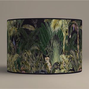 Può includere: Paralume cilindrico con una lussureggiante stampa della giungla. Il design presenta una fitta vegetazione verde, palme e un leopardo. Lo sfondo è scuro, con un bordo nero nella parte superiore e inferiore. Tema tropicale.