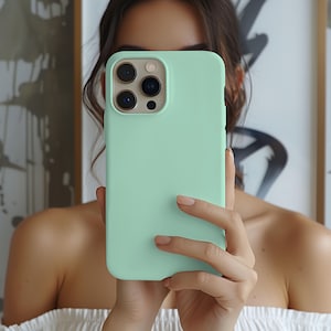 May include: A mint green phone case held in front of a person's face. The phone case has a smooth, matte finish and a camera cutout. The phone case is covering the phone's camera lenses.