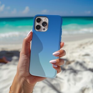 May include: A blue ombre phone case held in a hand. The case transitions from a deep blue at the top to a lighter blue at the bottom. The phone's camera lenses are visible. The background shows a beach with white sand and turquoise water.