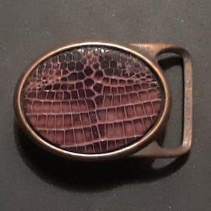 May include: A brown leather belt buckle with a textured, embossed design. The buckle is oval shaped and has a dark brown color.