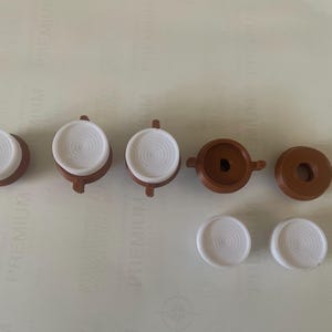 Puede incluir: Siete botones de plástico marrón y blanco con un centro elevado. Los botones son de forma circular y están dispuestos en fila.