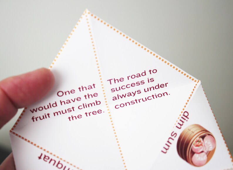 May include: White paper fortune cookie with a red and gold border. The fortune cookie contains the text "One that would have the fruit must climb the tree. The road to success is always under construction."