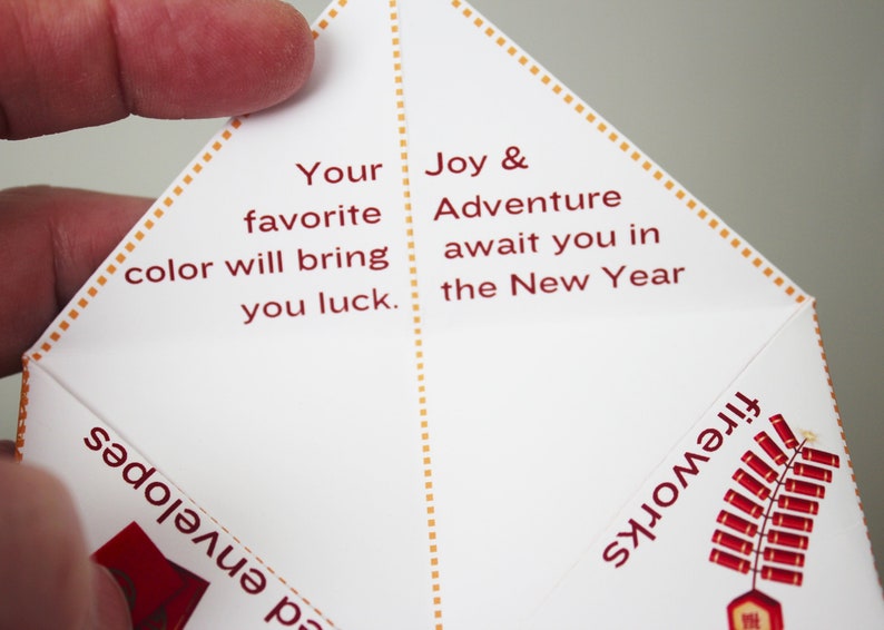 May include: A white paper fortune teller with red text. The text reads "Your favorite color will bring you luck. Joy & Adventure await you in the New Year." The fortune teller has a red and gold design on the outside.
