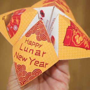 May include: A yellow paper fortune teller with red and white accents. The fortune teller has the words "Happy Lunar New Year" printed on one of the flaps.