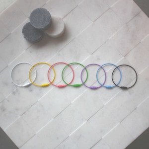 May include: Seven colorful key rings in a row on a white tiled surface. The key rings are white, yellow, pink, green, light blue, purple, and black.