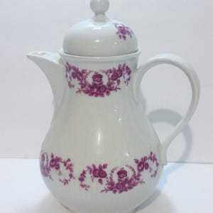May include: White ceramic coffee pot with a lid and handle. The pot is decorated with a floral pattern in shades of purple. The design is repeated around the body and on the lid of the pot.