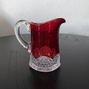 以下が含まれることがあります： ルビーレッドのボディと透明なガラス製のハンドルが付いた、小さな透明なガラス製のクリーマー。クリーマーは、ベースの周りにカットガラスのデザインが施されています。