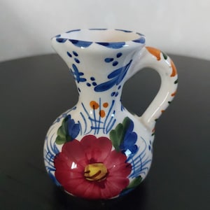 May include: A small, white ceramic pitcher with a rounded body and a handle. The pitcher is decorated with a large red flower, blue and green leaves, and blue and orange accents. The handle has orange and green details.