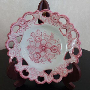 以下が含まれることがあります： ピンクの花柄とスカラップエッジが特徴の白い装飾皿。エッジにはハート、三角形、円などの切り抜き形状があります。お皿は濃い茶色の木製スタンドに飾られています。