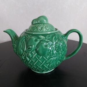Peut inclure: Une théière en céramique verte avec un couvercle orné d'un motif en relief de fruits et de feuilles. La théière a un corps rond et une poignée sur le côté.