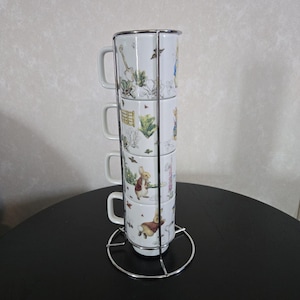 以下が含まれることがあります： ピーターラビットのデザインが施された白いセラミック製のマグカップ4個セットが、シルバーの金属製スタンドに積み重ねられています。マグカップには、ピーターラビットと本に登場する他のキャラクターの異なるイラストが描かれています。