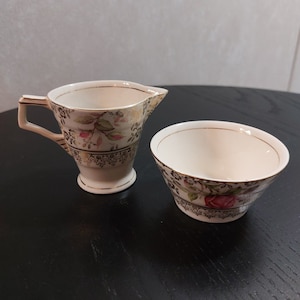 May include: A cream-coloured creamer and sugar bowl set. The set has a floral design with pink and green accents, gold trim, and a decorative black border. The creamer has a handle and spout.