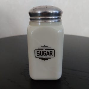 May include: A vintage white glass sugar shaker with a silver metal lid. The word "SUGAR" is printed in a black rectangular frame on the front. The shaker is rectangular with rounded corners.