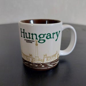 May include: White ceramic mug with the word "Hungary" in green lettering. The mug has a brown interior and a detailed illustration of a landmark. The Starbucks Coffee logo is also visible.