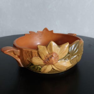 Puede incluir: Un cuenco de cerámica marrón con un diseño floral. El cuenco tiene dos asas y está decorado con una flor grande y hojas.