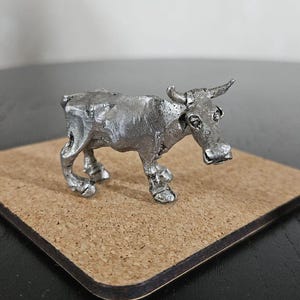 Peut inclure: Petite figurine de vache argentée avec des cornes et des détails. La vache est posée sur un dessous de verre carré en liège. La figurine a un aspect rustique et artisanal, probablement un objet décoratif.