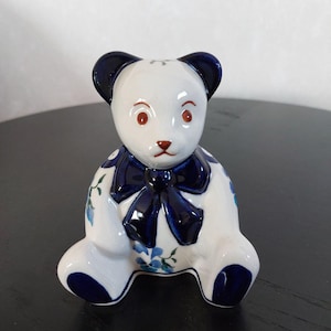 Puede incluir: Figura de oso de peluche de cerámica con cuerpo blanco, orejas azul marino y lazo a juego. El oso tiene ojos rojos y una pequeña nariz roja. Acentos florales azules decoran el cuerpo. El oso está sentado.