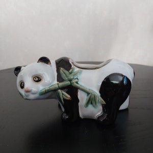 Puede incluir: Una figurita de panda de cerámica con pelaje blanco y negro, sosteniendo un tallo de bambú verde en su boca. La figurita está esmaltada en blanco y negro con acentos verdes.