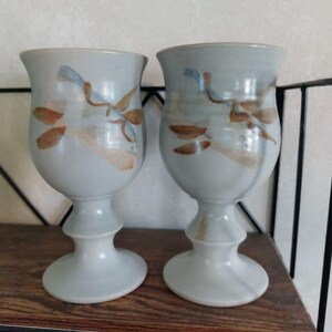 Könnte beinhalten: Zwei Keramikbecher mit hellblauer Glasur und braunen und blauen floralen Designs. Die Becher haben eine abgerundete Schalenform und einen Sockel. Die Becher werden auf einem Holzregal ausgestellt.