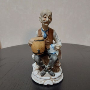May include: A ceramic figurine of a man with a white beard and moustache, wearing a brown waistcoat and a blue tie, holding a barrel and a mug. He is sitting on a white base with a brown and white design.