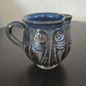 May include: A blue ceramic creamer with a textured design of stylized fish. The creamer has a handle and a rounded body.