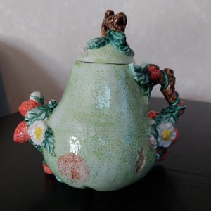 Puede incluir: Una tetera verde en forma de pera con un oso marrón en la tapa y un asa hecha de una rama marrón con fresas y hojas. La tetera está decorada con fresas, hojas y flores blancas.