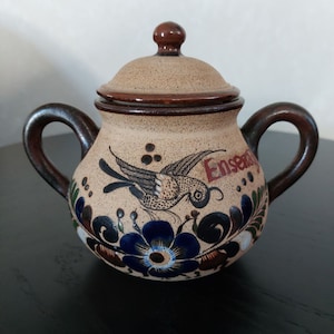 May include: A brown ceramic sugar bowl with a lid, featuring a painted design of a bird with blue and green floral accents. The bowl has a handle on each side and the word "Ensenada" is painted on the side.