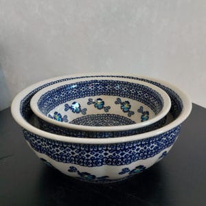 May include: Two ceramic mixing bowls nested together. The bowls are white with a blue floral pattern and a decorative blue border. The floral pattern includes small blue flowers. The bowls are likely for kitchen use.