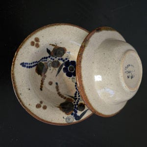 May include: A beige ceramic bowl and saucer set with a floral design in brown and blue. The rim of the bowl and saucer is brown. The saucer has a pattern of brown dots and blue lines. The bowl has text on the bottom.