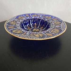 May include: A deep blue decorative bowl with a gold rim and ornate gold detailing. The bowl features a scalloped edge and a detailed pattern of gold figures and designs around the rim, creating an elegant aesthetic.