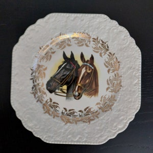 以下が含まれることがあります： 金色の縁取りと浮き彫りのデザインが施された白いセラミック製の正方形のプレート。プレートの中央には、黄色い背景に黒い馬と茶色の馬の頭が描かれた絵があります。