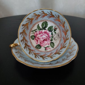 May include: A vintage teacup and saucer set in light blue with gold detailing. The teacup has a central floral design of a pink rose and a white rose. The rims are adorned with gold leaf patterns.