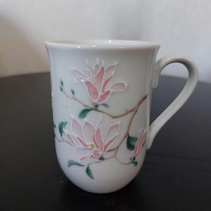 Pode incluir: Caneca de cerâmica branca com uma alça curva, adornada com flores de magnólia rosa e folhas verdes em galhos marrons. O design floral é delicadamente pintado na superfície da caneca, criando uma estética serena e elegante. Uma adição perfeita para qualquer cozinha ou conjunto de chá.