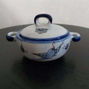 Puede incluir: Un cuenco pequeño de cerámica blanca con tapa, con diseños de pájaros y mariposas pintados a mano en azul. El cuenco tiene dos asas pequeñas y un asa de bucle en la tapa. El borde y las asas están pintados de azul.